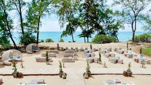 Un espacio al aire libre frente a la playa con varias mesas y sillas de mimbre, rodeado de plantas en macetas y árboles.