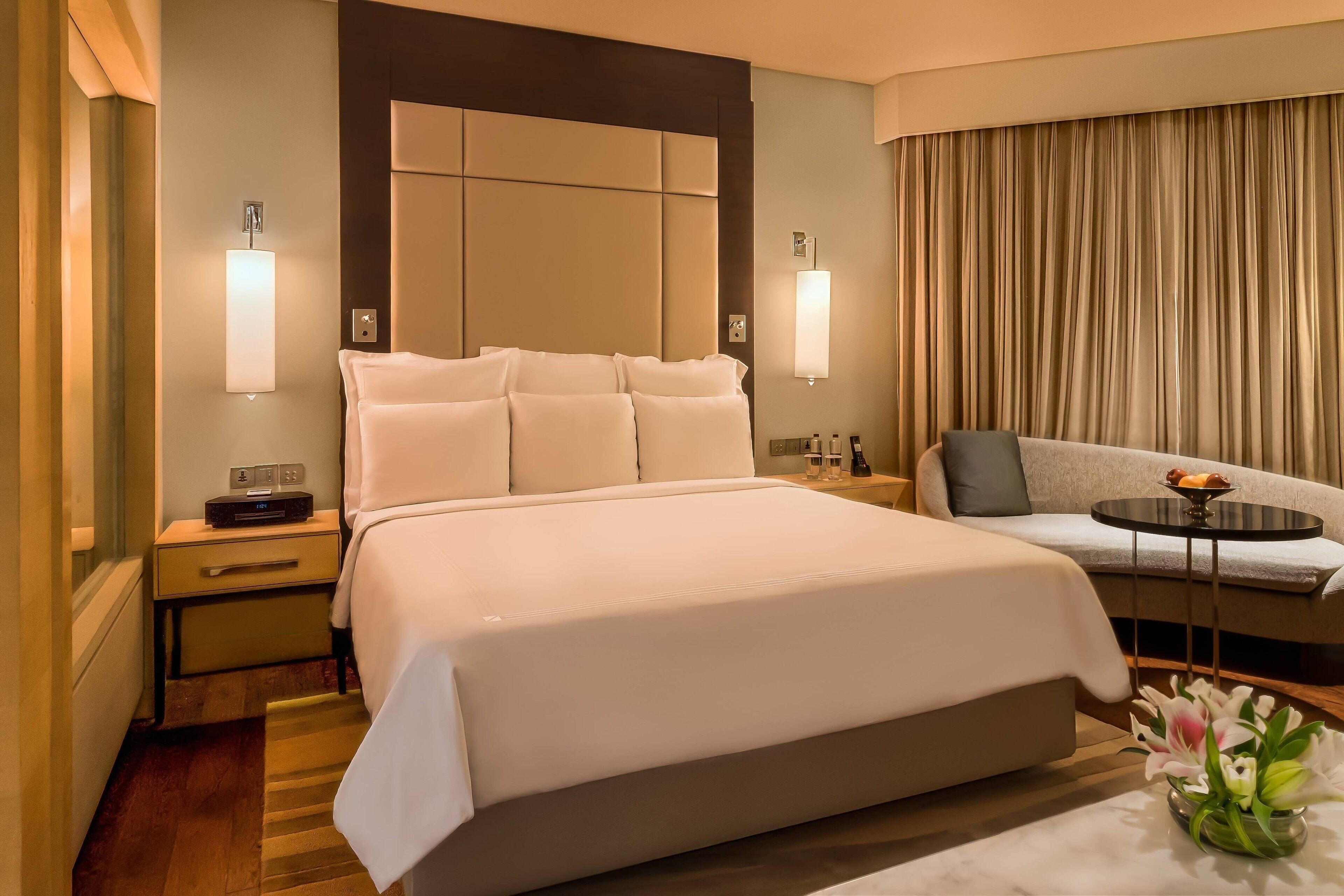 Habitación de hotel con una cama grande, un sofá, una mesita con frutero y un florero con flores.