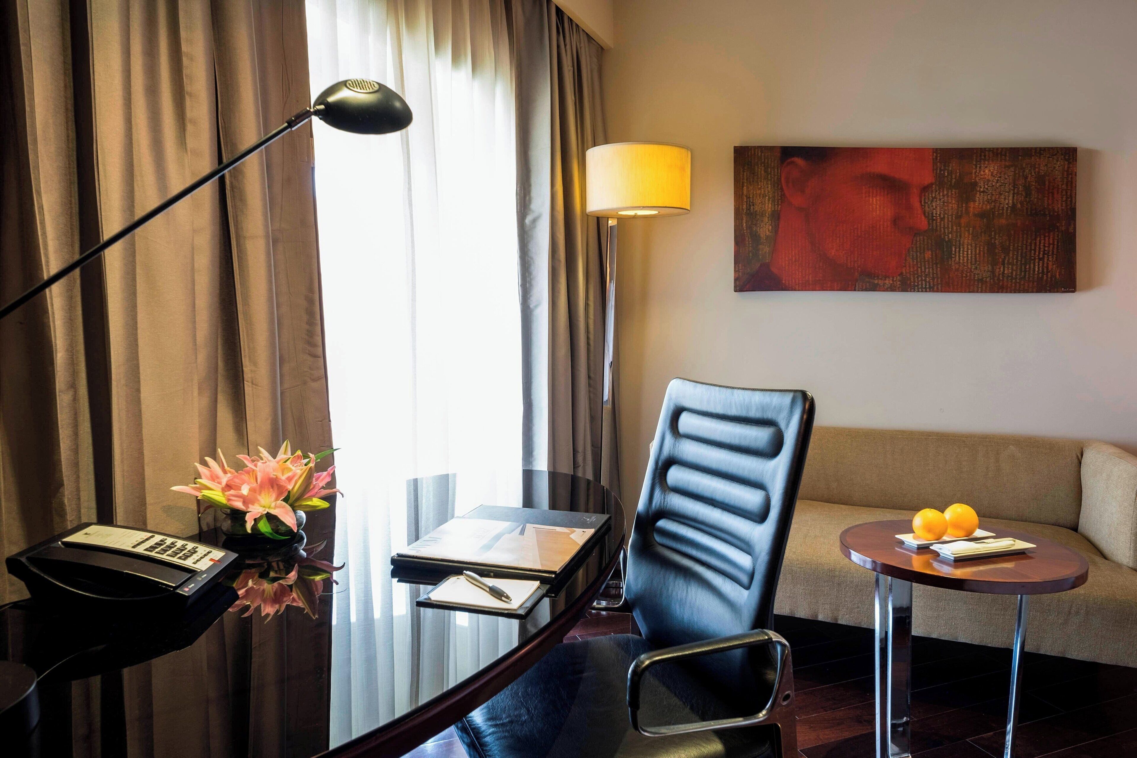 Habitación de hotel con escritorio, silla, lámpara, sofá y una pequeña mesa con fruta.