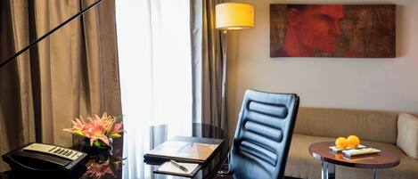 Habitación de hotel con escritorio, silla, lámpara, sofá y una pequeña mesa con fruta.