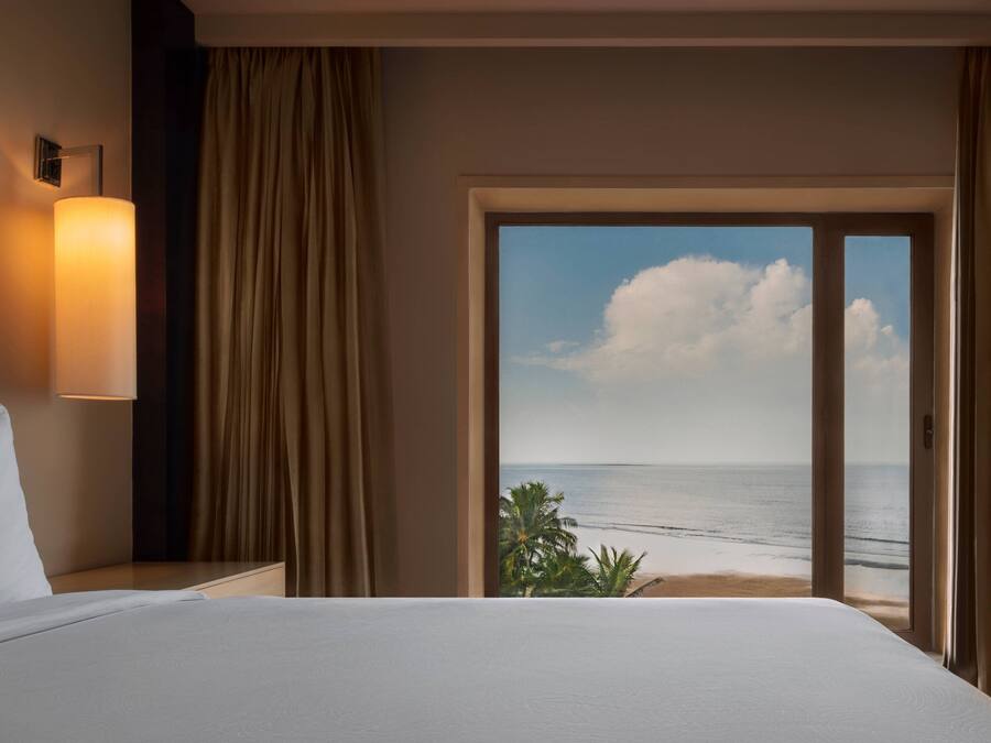 Quarto de hotel com uma cama, mesa de cabeceira, luminária e vista para o oceano através de uma grande janela.