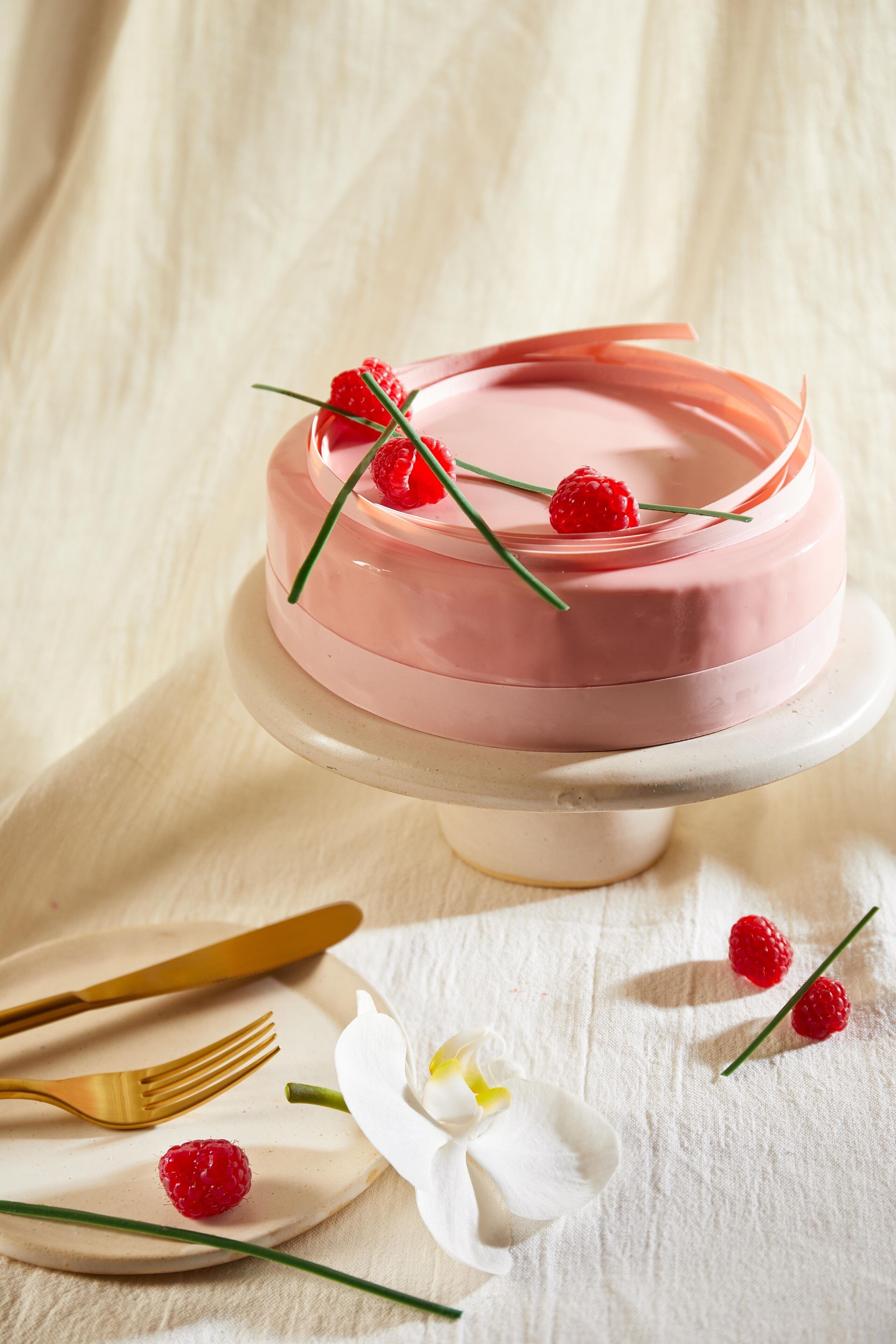 Un gâteau aux framboises, présenté sur une assiette blanche, accompagné d’une fourchette et d’un couteau, et d’une fleur d’orchidée.