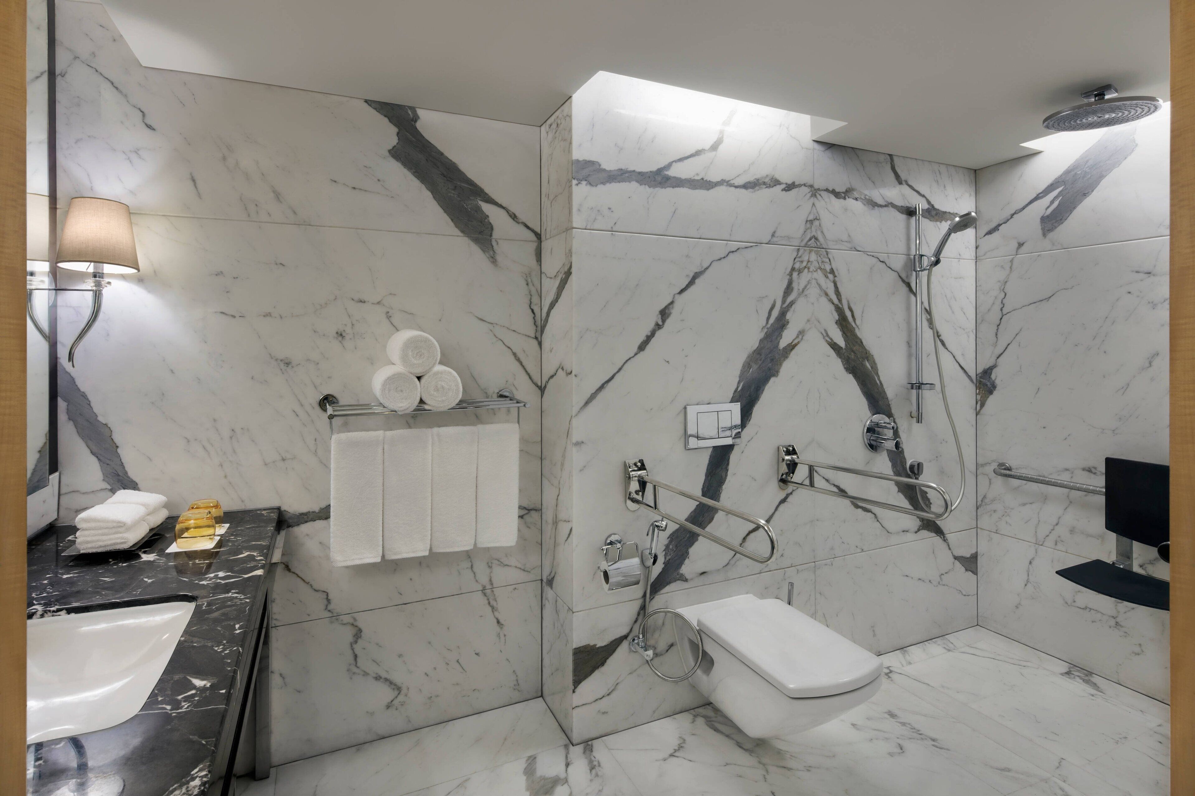 A bathroom with a marble floor and walls, a white toilet, a sink, a shower with a handheld showerhead, and a towel rack with towels.