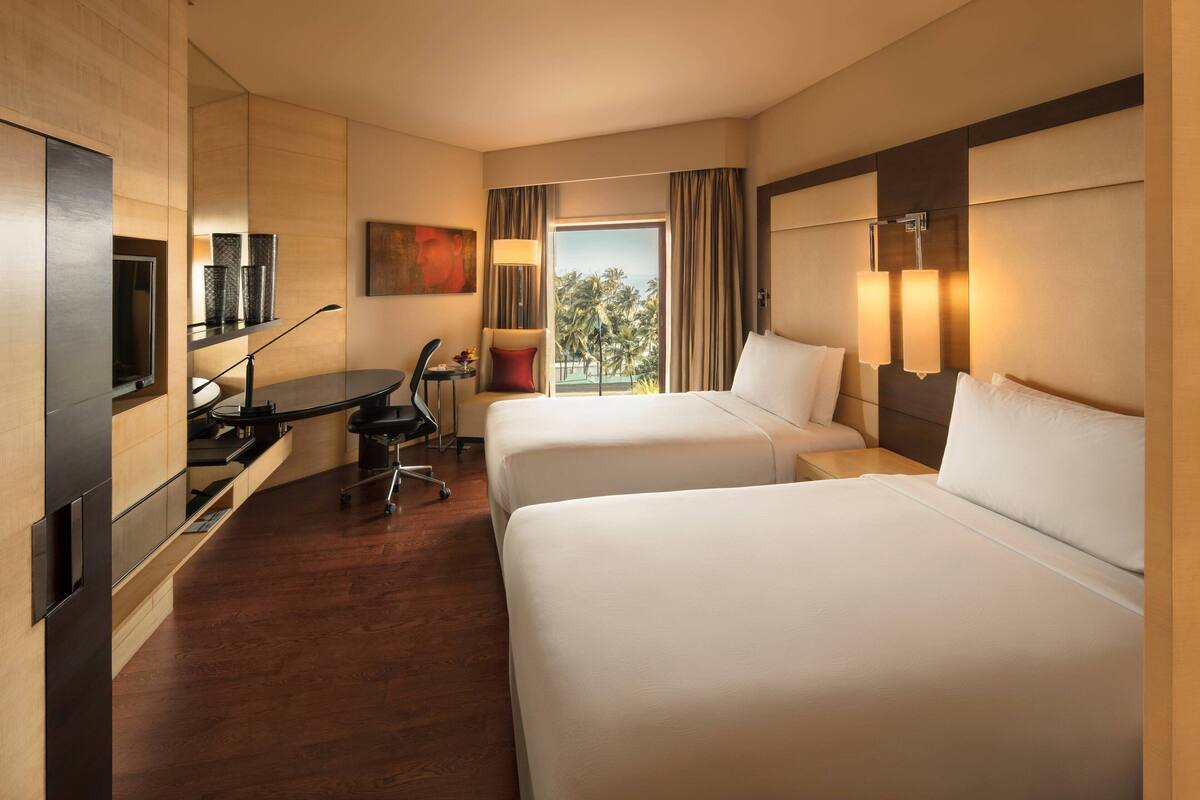 Ein Hotelzimmer mit zwei Betten, einem Schreibtisch, einem Stuhl, einem Fernseher und einem Fenster mit Blick auf Palmen.