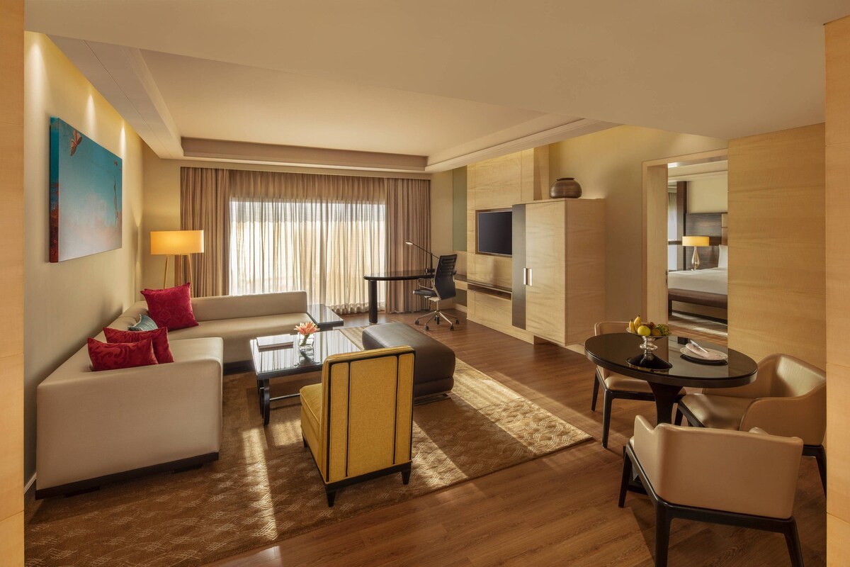 A modern hotel room with a sofa, armchair, dining table, and a TV.