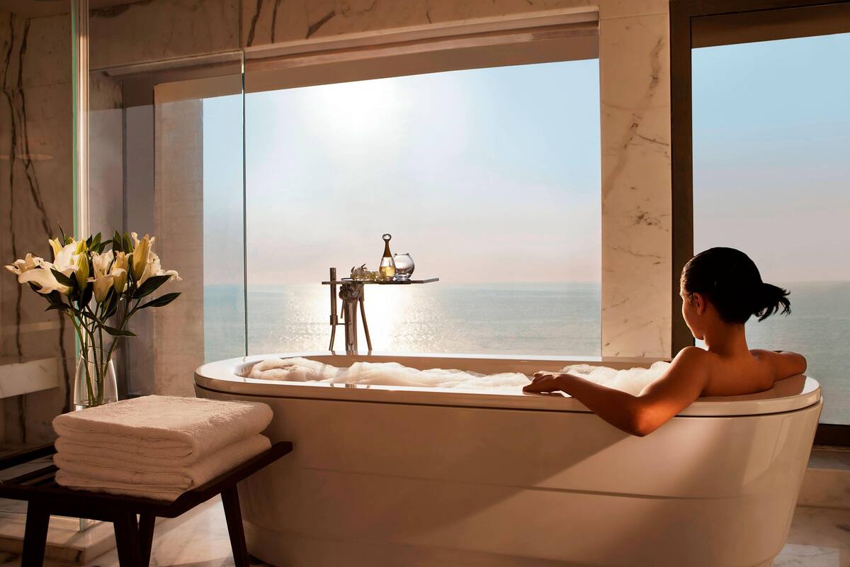 Eine Frau entspannt in einer Badewanne mit Blick auf das Meer. Ein Tisch mit Blumenvase und Tablett mit Pflegeprodukten steht daneben.