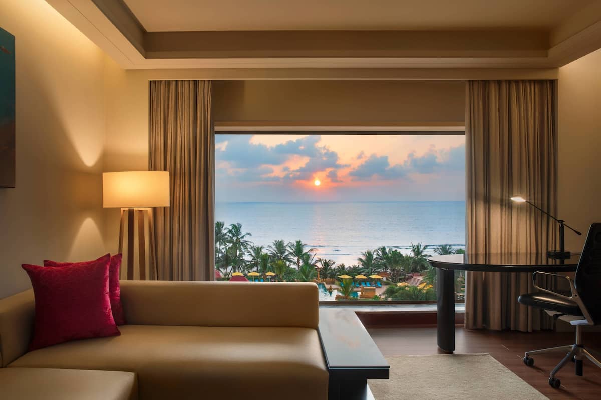 Ein Hotelzimmer mit einem großen Fenster, das einen Blick auf das Meer und Palmen bei Sonnenuntergang bietet.