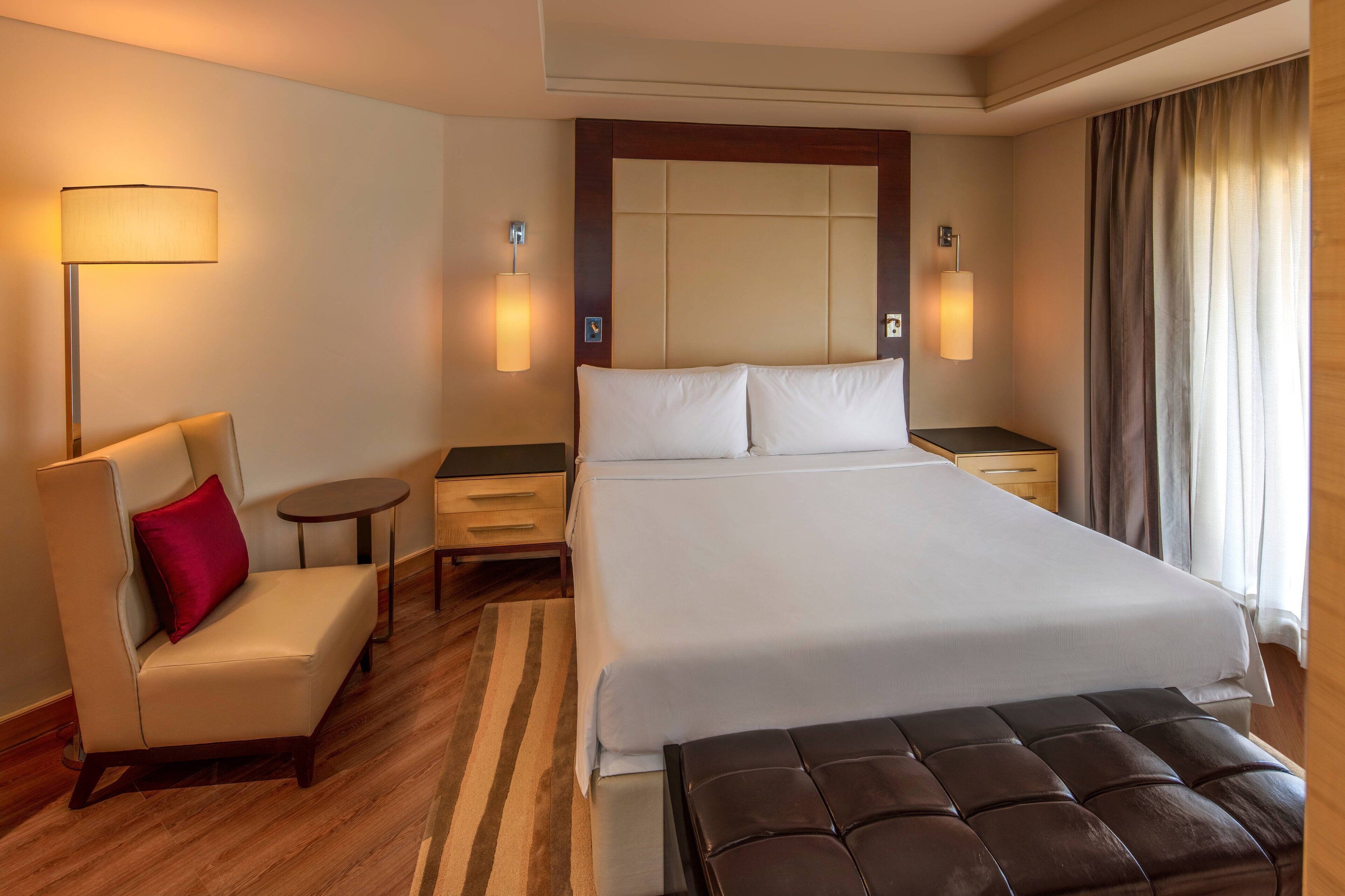 A hotel room with a large bed, a bedside table, a chair, a lamp, and a window with curtains.