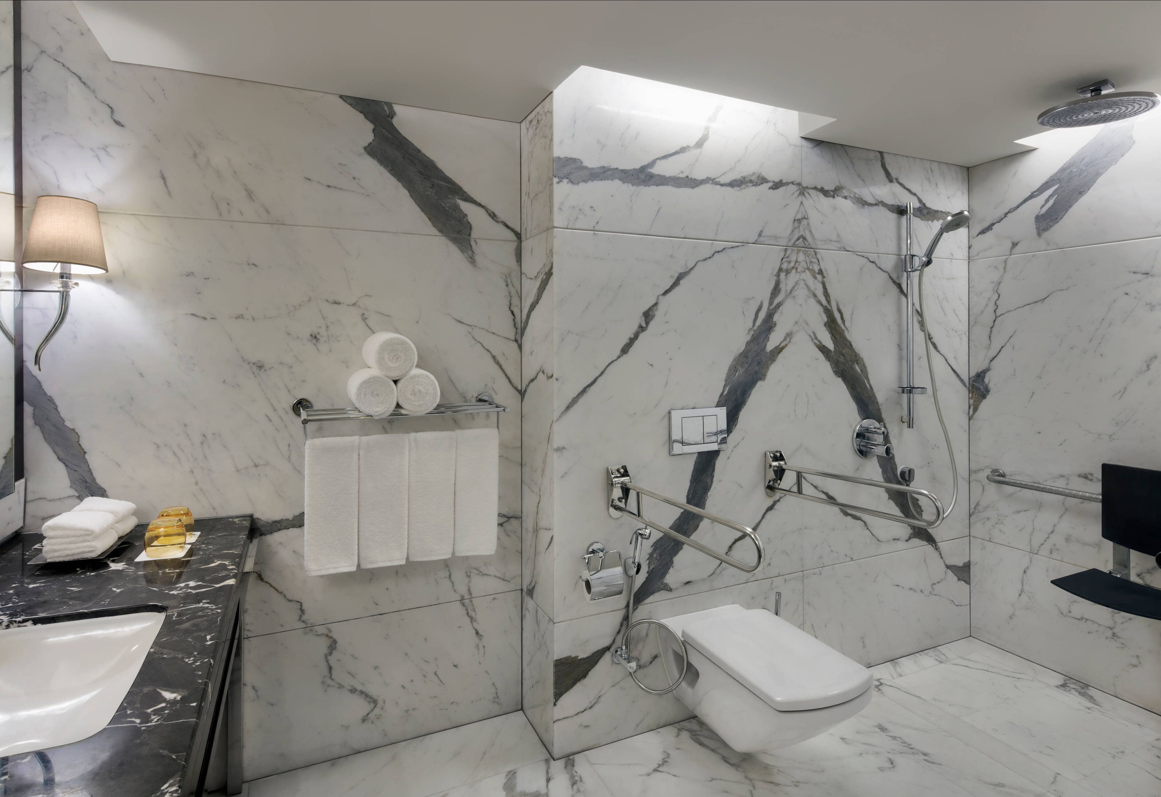 Un baño con piso y paredes de mármol, un lavamanos blanco, un inodoro y una zona de ducha con barras de apoyo.