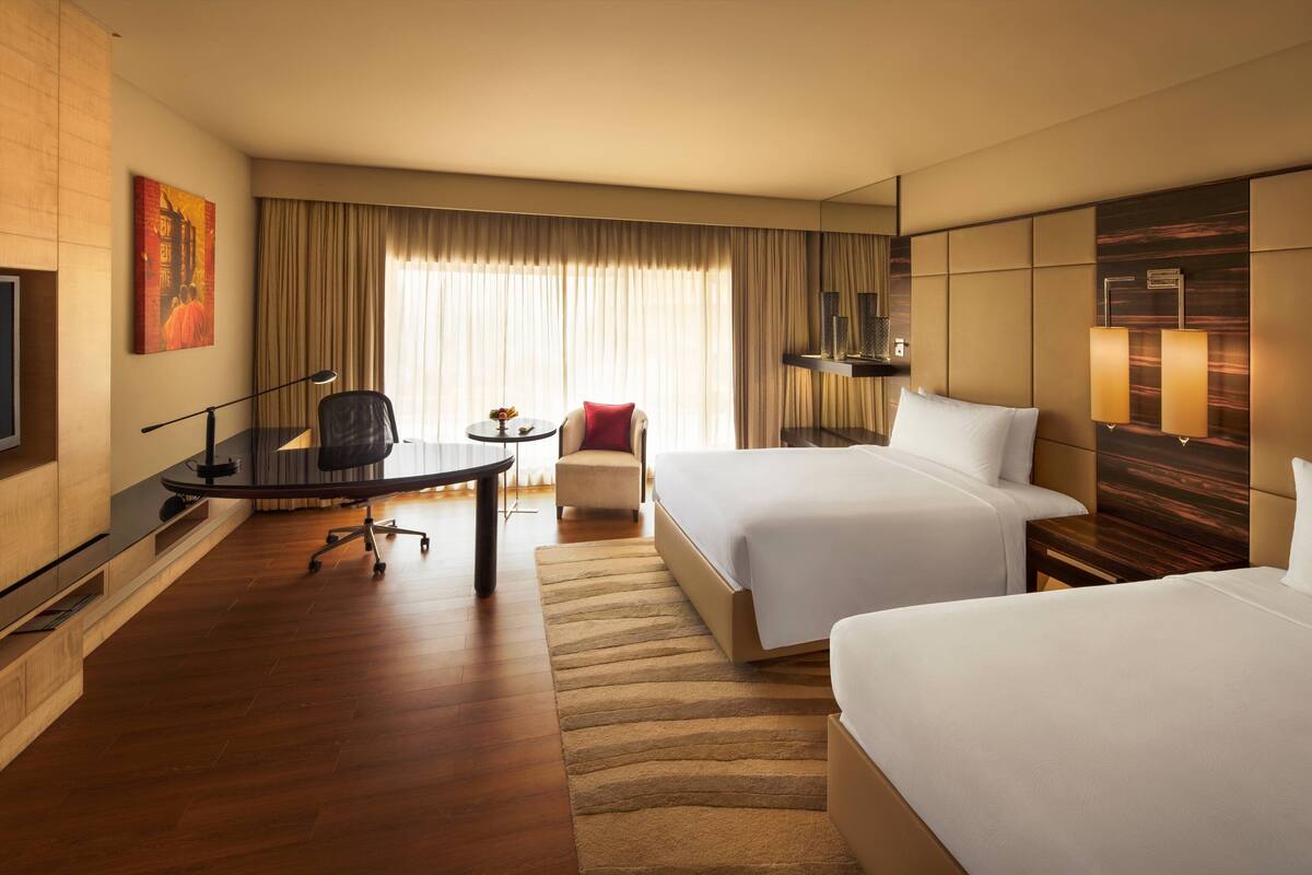 Ein Hotelzimmer mit zwei Betten, einem Schreibtisch, einem Sessel, einem Fernseher und einem großen Fenster mit Vorhängen.