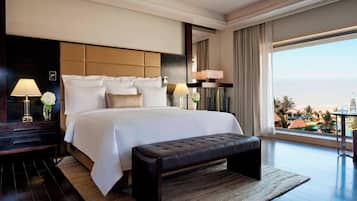 A modern hotel room with a large bed, a bench, two bedside tables with lamps, and a view of the ocean.