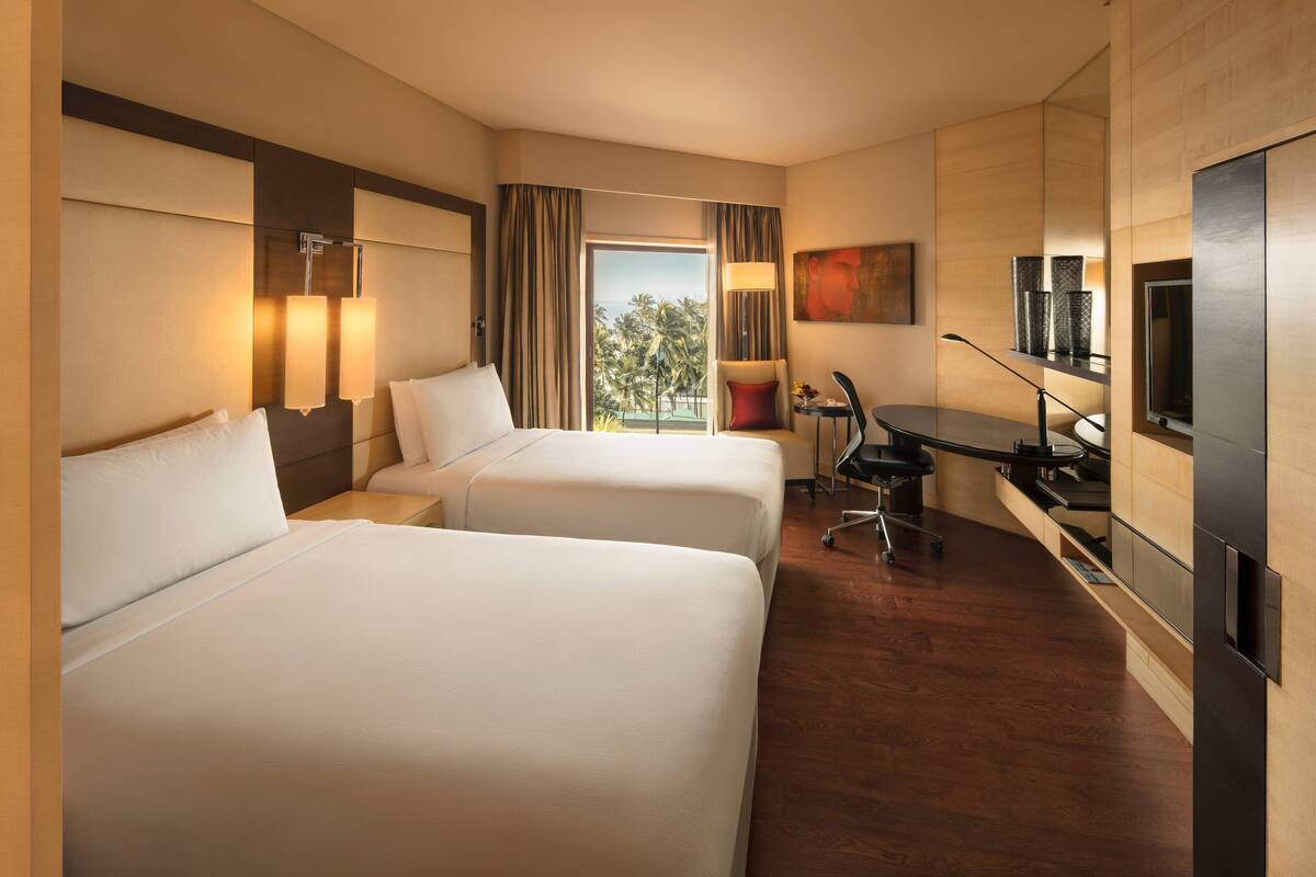 Ein Hotelzimmer mit zwei Betten, einem Schreibtisch, einem Stuhl, einem Fernseher und Blick auf Palmen.