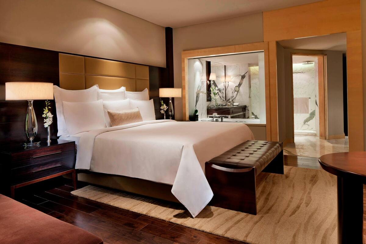 Ein modernes Hotelzimmer mit einem großen Bett, Nachttischlampen, einer Bank und Blick auf das Badezimmer durch eine Glastür.