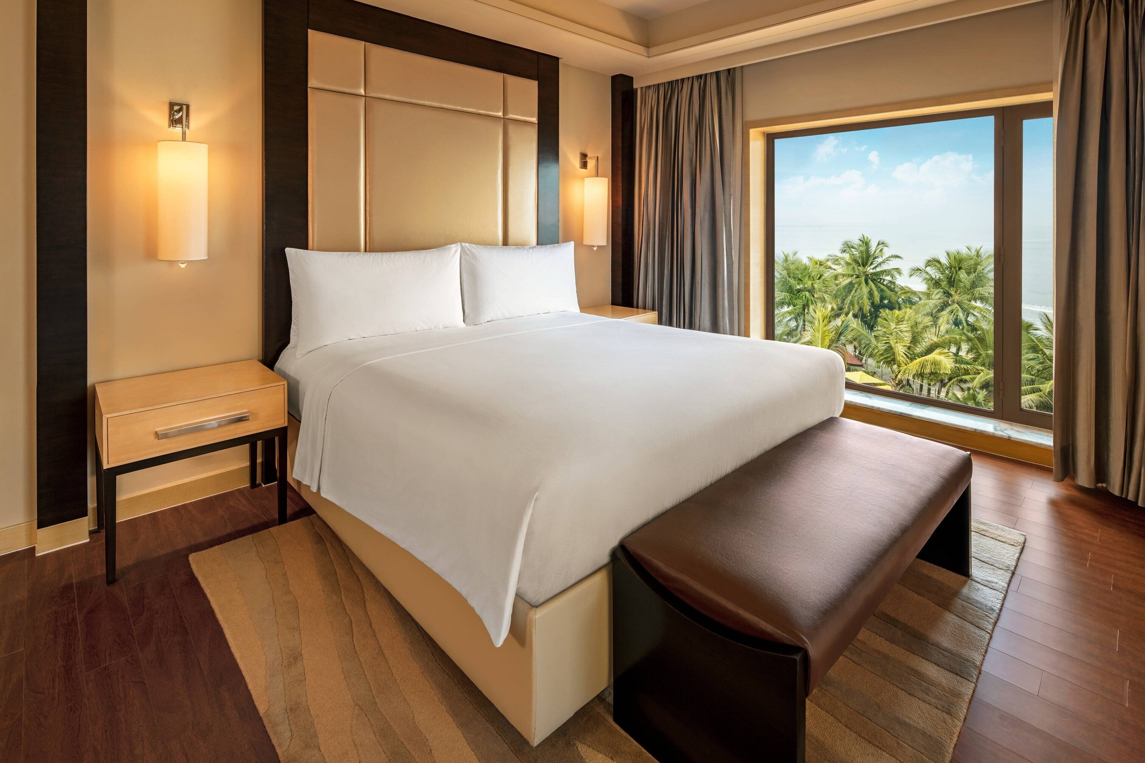Habitación de hotel con una cama grande, un banco, una mesita de noche y vista a palmeras a través de una ventana.