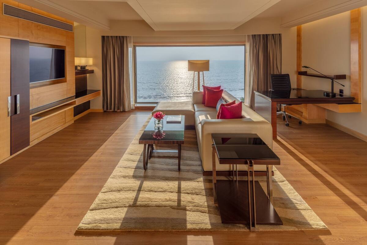 Ein modernes Hotelzimmer mit großem Fernseher, einer Couch mit roten Kissen, einem kleinen Tisch mit einer Vase und Blick aufs Meer.