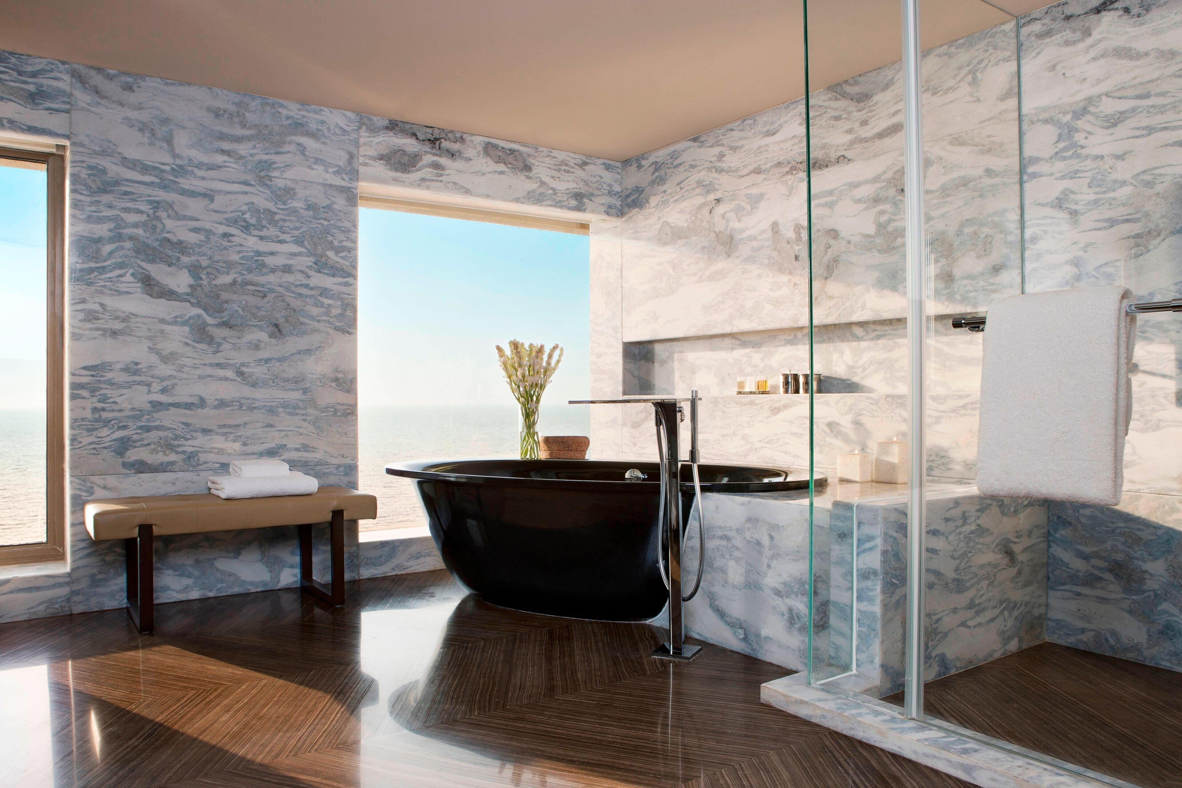 A modern bathroom with a freestanding bathtub, marble walls, and a bench by the window.