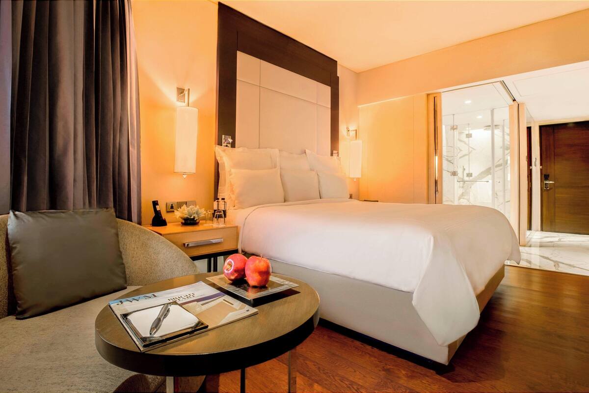 Ein Hotelzimmer mit einem großen Bett, einem Nachttisch mit Lampe, einem Sofa und einem kleinen runden Tisch mit Zeitschrift und Stift.