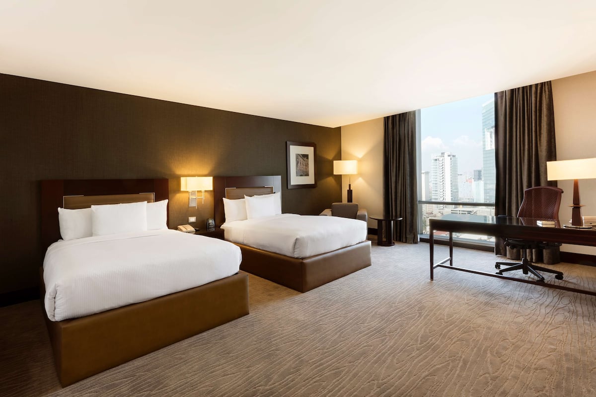 Habitació d'hotel amb dos llits, una escriptori, una cadira i una finestra gran amb vista a la ciutat.