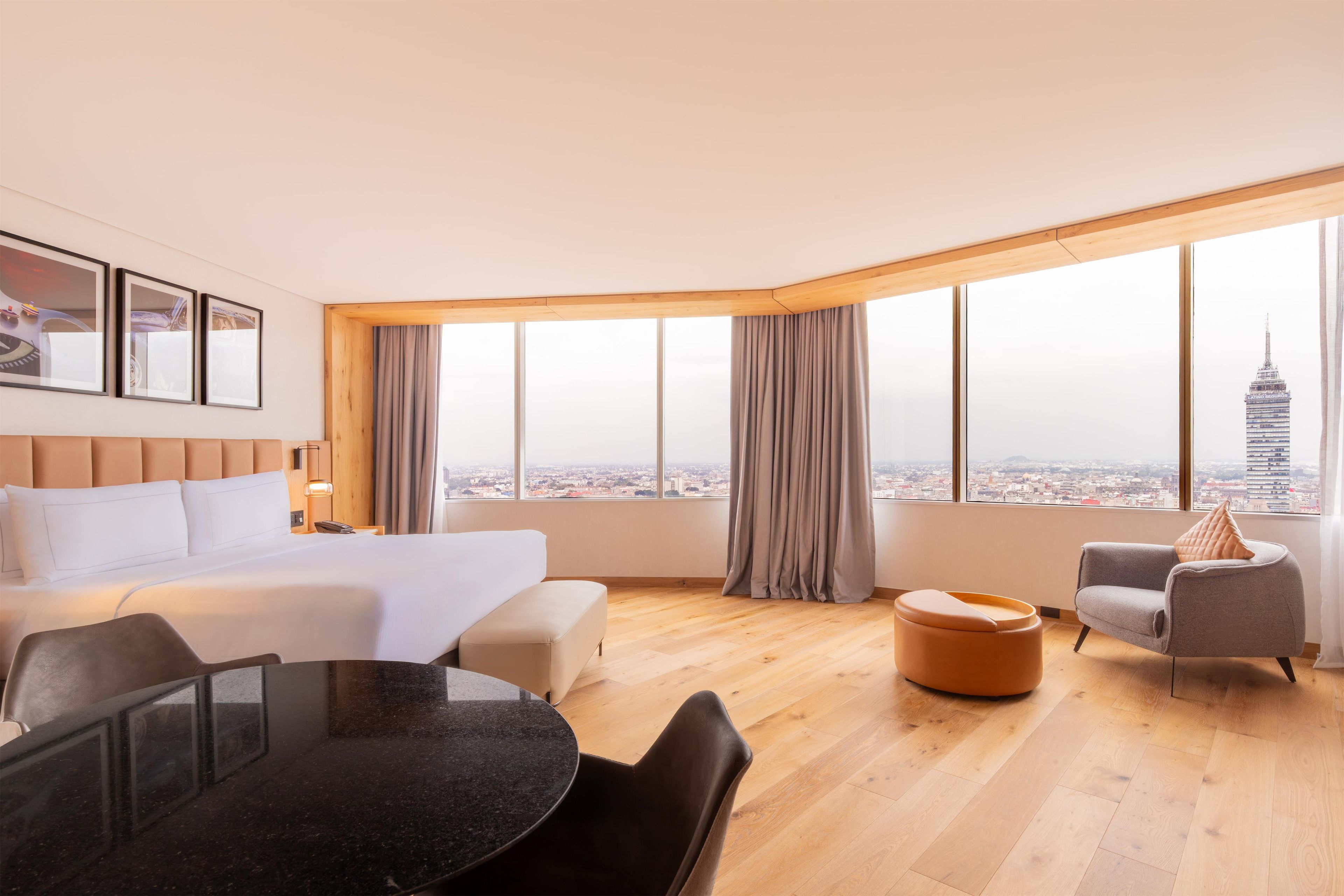 Habitació d'hotel moderna amb un llit gran, una taula de menjador i vista a un paisatge urbà a través de grans finestres.