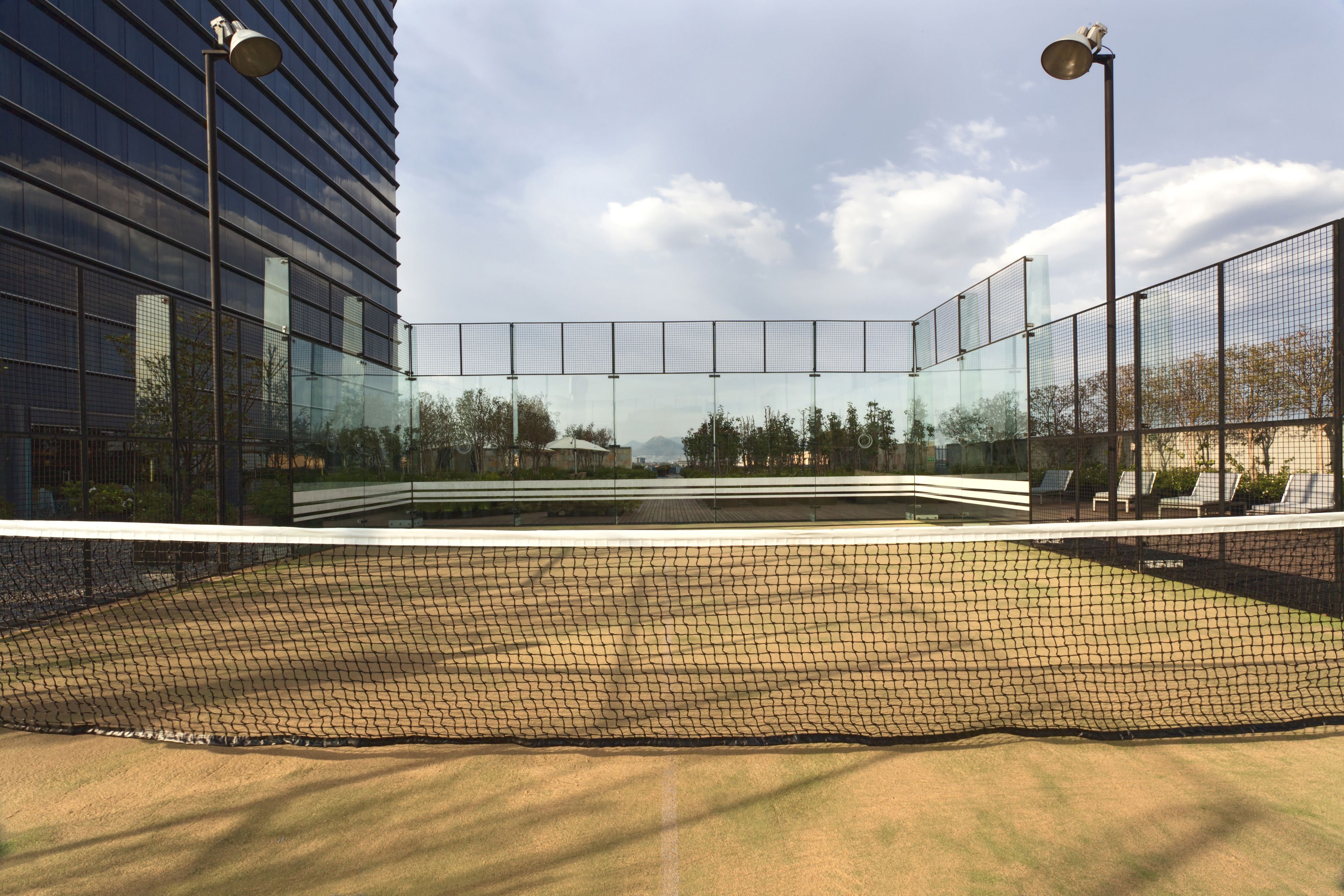 A tennis court with a net, surrounded by a fence, and adjacent to a modern building with glass windows.