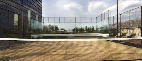 A tennis court with a net, surrounded by a fence, and adjacent to a modern building with glass windows.