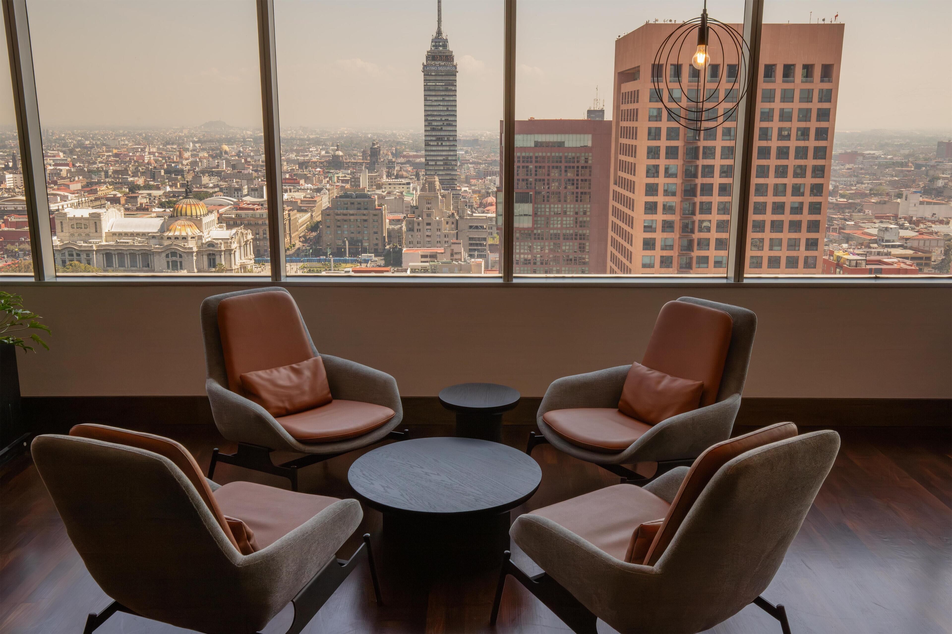 A room with a large window offering a city view, two armchairs, a small round table, and a hanging light fixture.