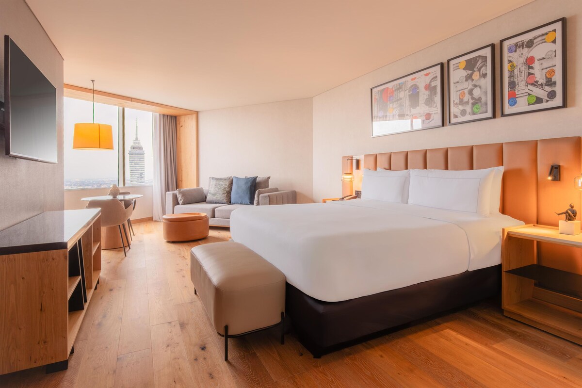 Habitación de hotel con una cama grande, un escritorio, una silla, un sofá y vistas de un edificio alto.