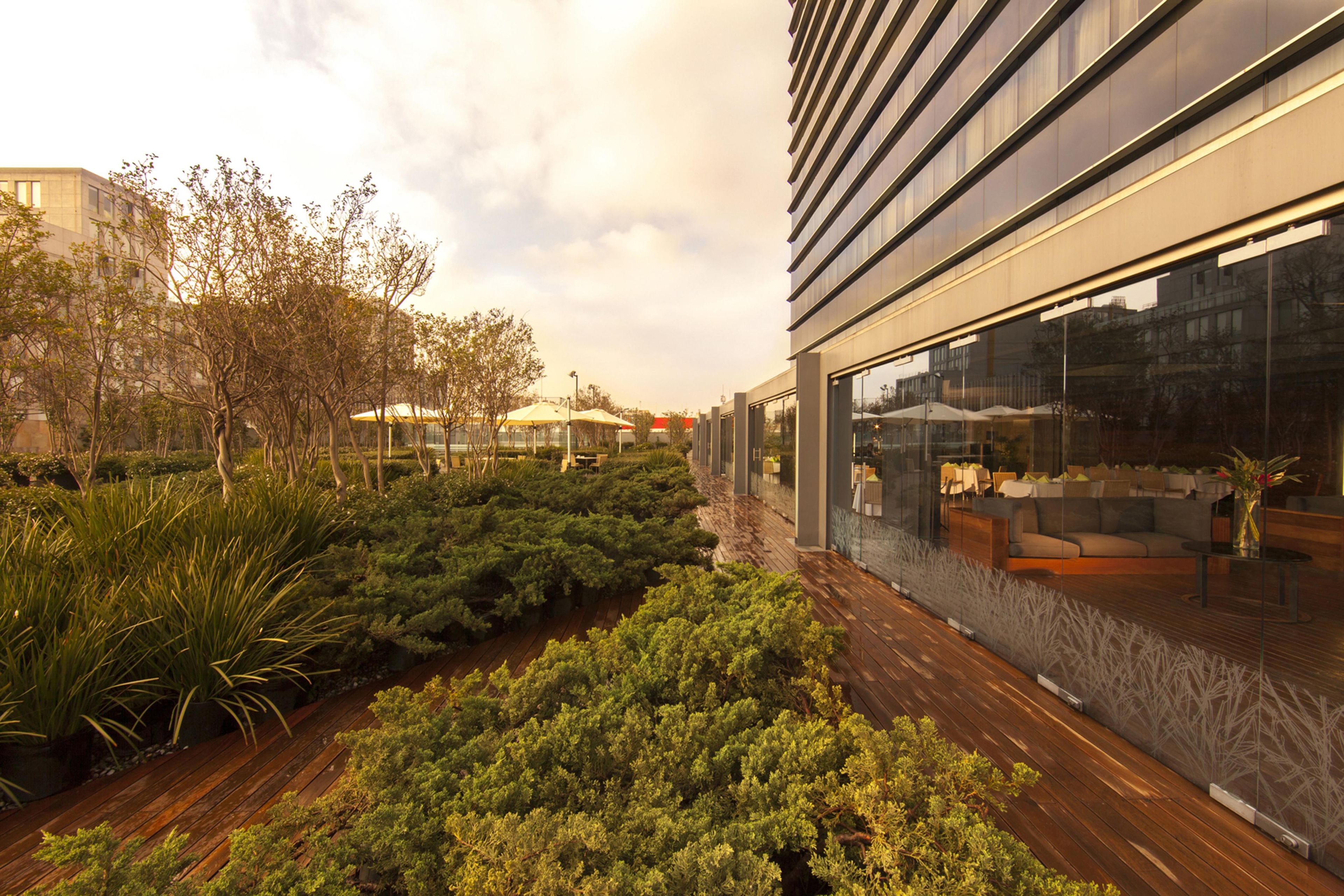 Un edificio moderno con fachada de vidrio, terraza de madera y jardín ajardinado con arbustos y árboles variados.