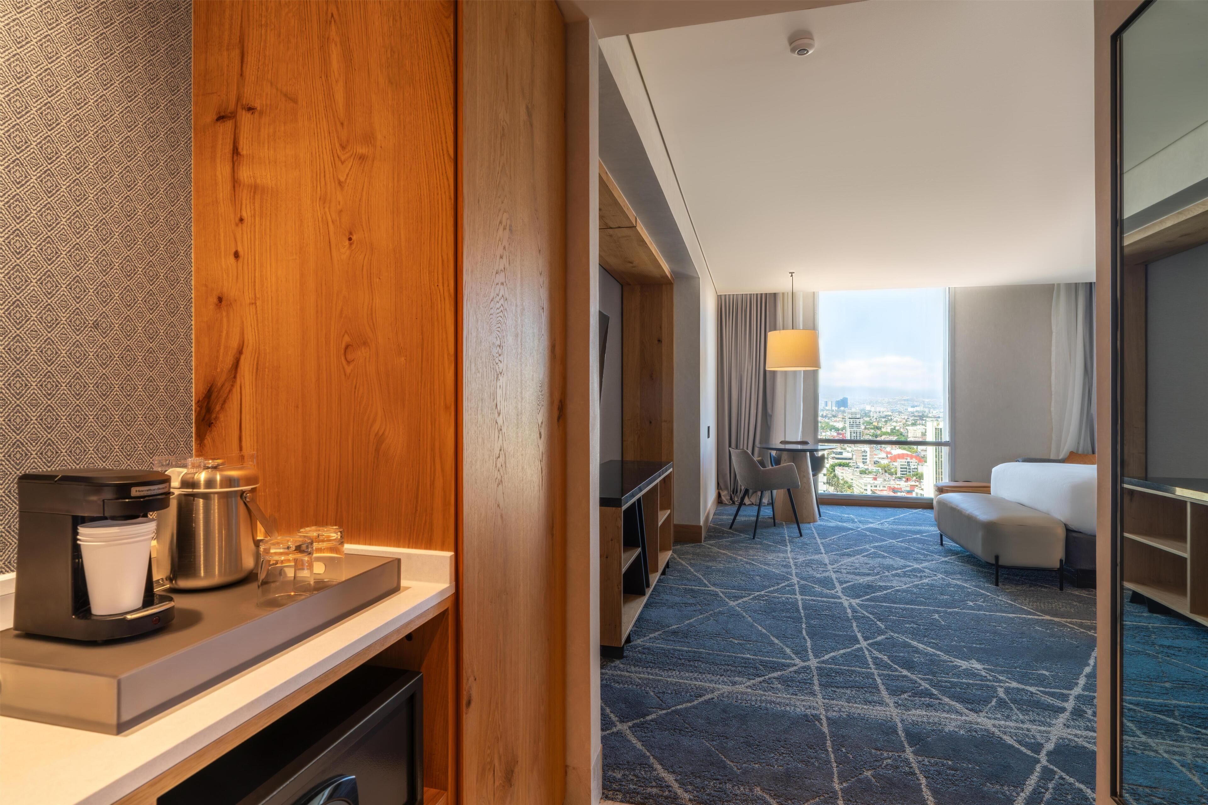 A hotel room with a wooden accent wall, a coffee machine, a dining area with a view, and a sofa.