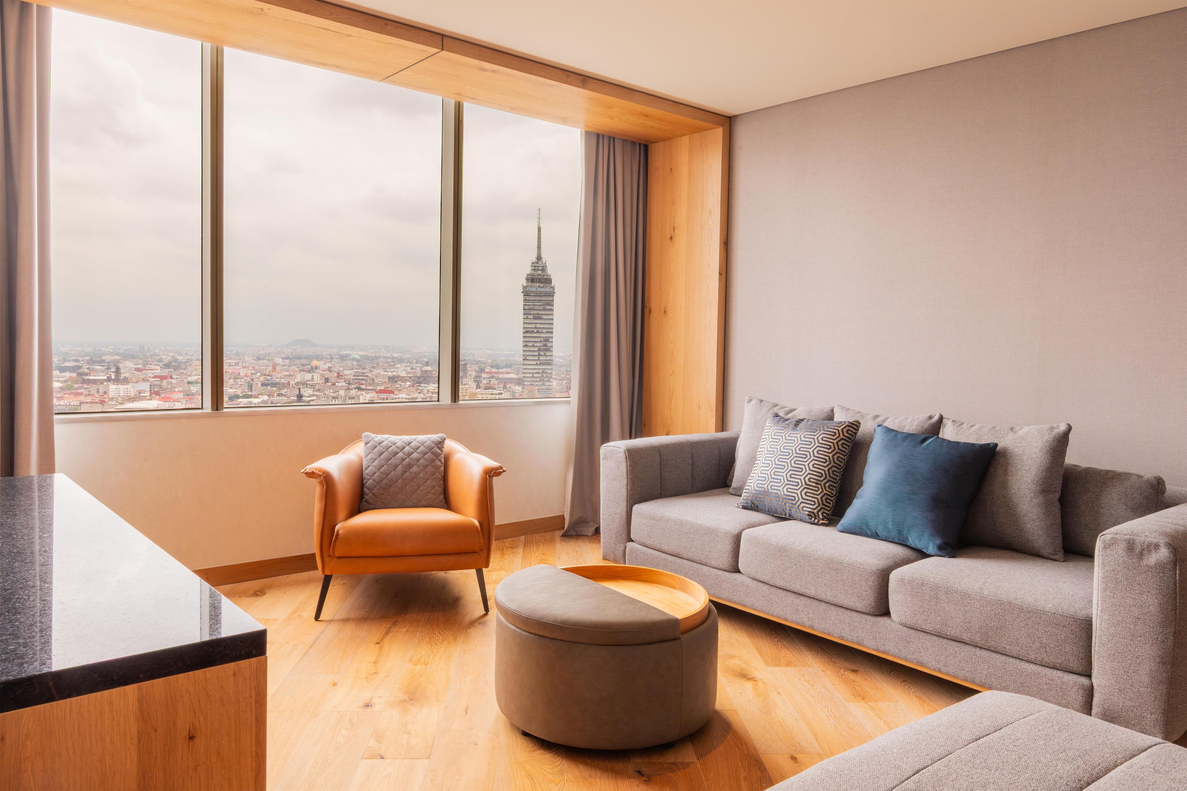 Ein modernes Wohnzimmer mit Sofa, Hocker und Sesseln, großen Fenstern mit Blick auf die Stadt und Holzboden.