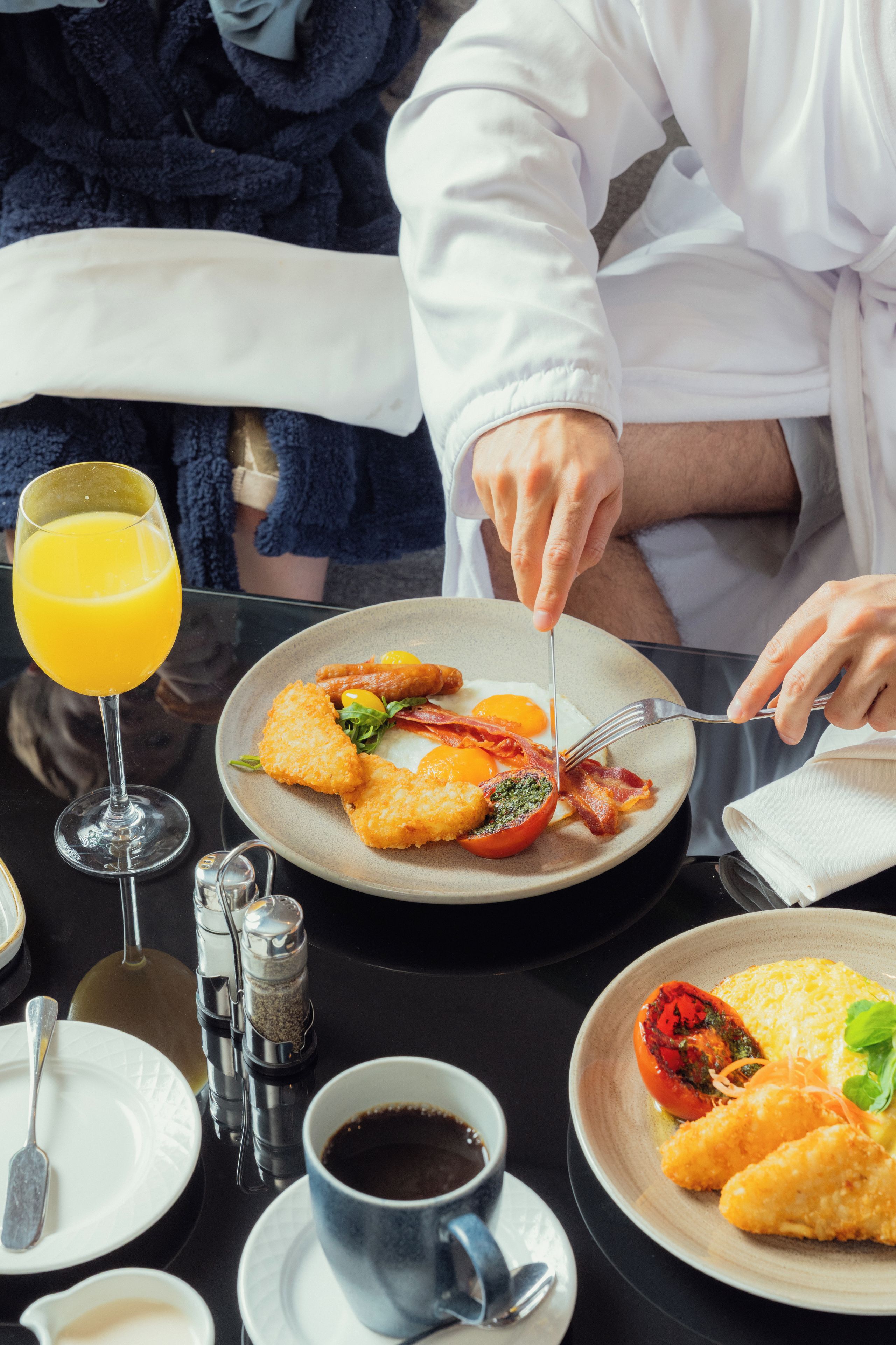 En morgenmadsbordopstilling med en tallerken med æg, bacon og en tomat, et glas appelsinjuice og en kop kaffe.