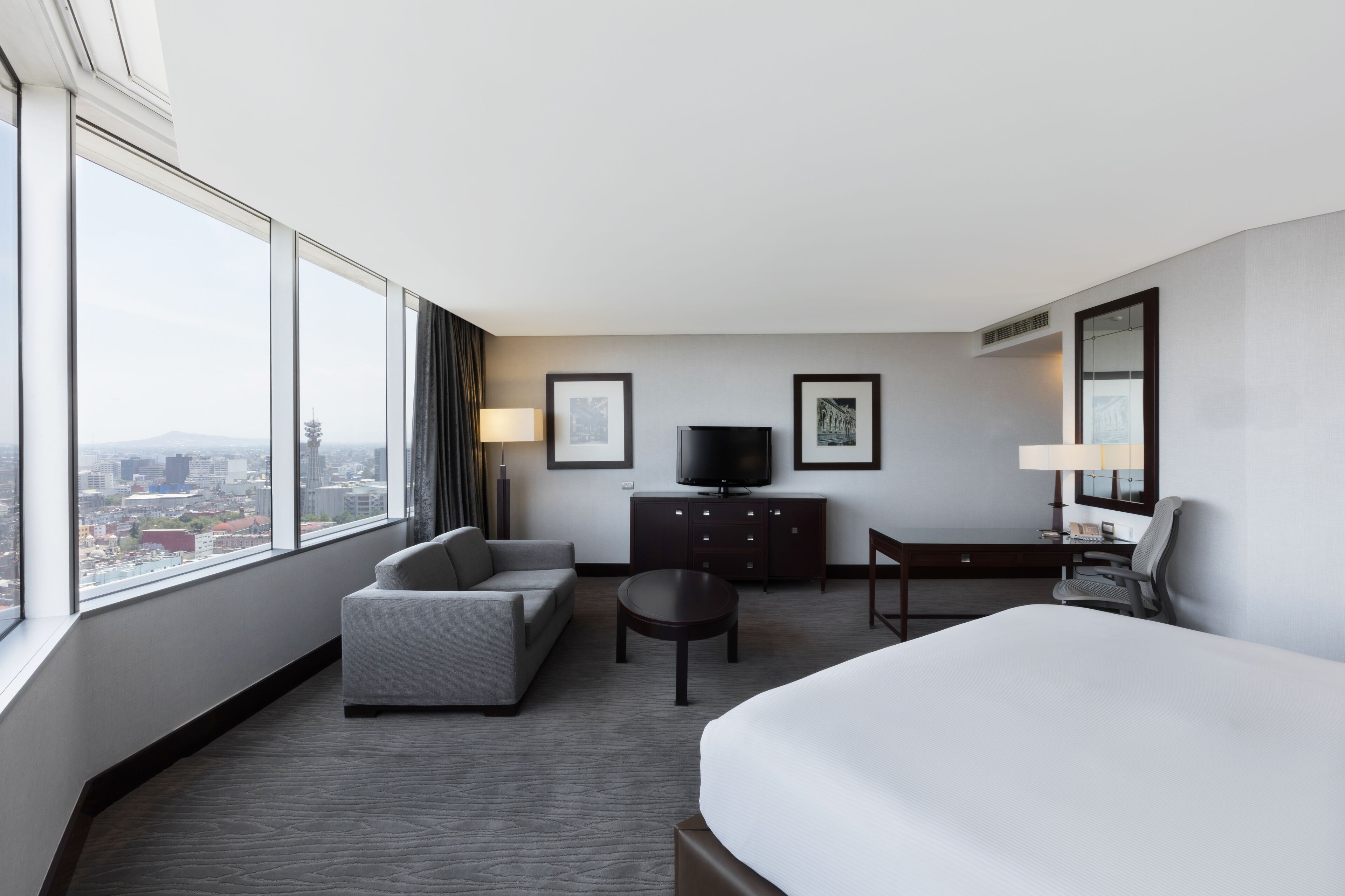 Une chambre d’hôtel avec un grand lit, un canapé, un bureau et une télévision. De grandes fenêtres offrent une vue sur la ville.