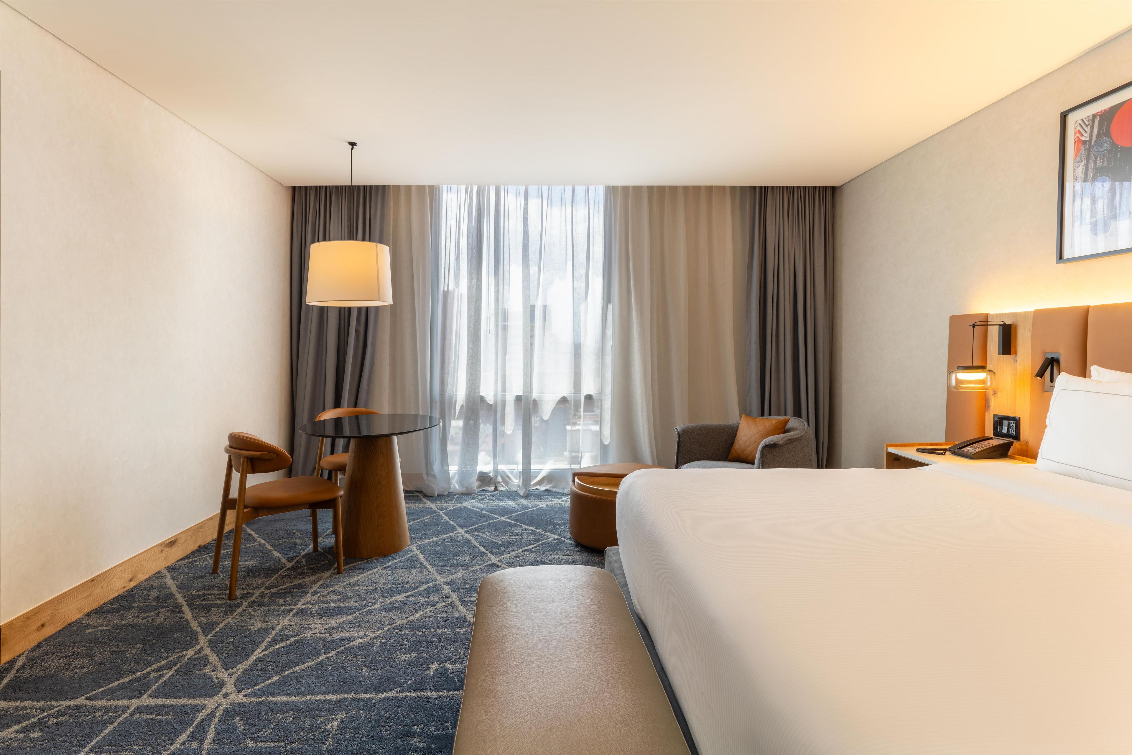 A hotel room with a large bed, a desk with a chair, a small table, a sofa, and a window with curtains.