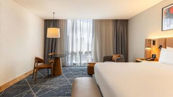A hotel room with a large bed, a desk with a chair, a small table, a sofa, and a window with curtains.