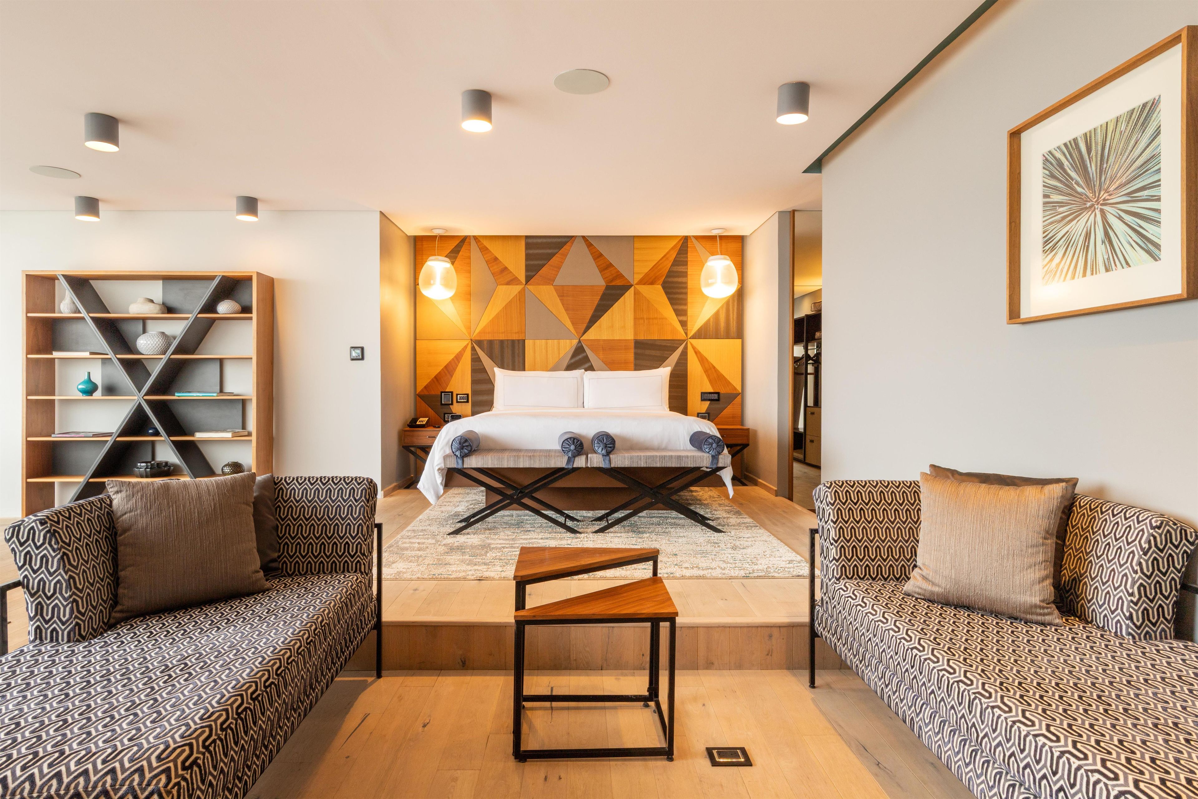 Ein modernes Schlafzimmer mit einem Bett, zwei Sofas, einem kleinen Tisch und einer Wand mit geometrischem Muster.