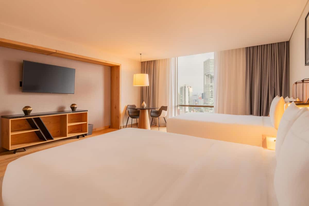 Habitació d'hotel amb un llit gran, un televisor fixat a la paret, una taula consola de fusta, un escriptori amb dues cadires i vista a la ciutat a través de grans finestres.