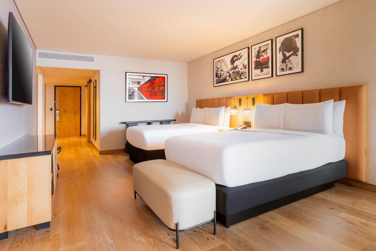 Habitació d'hotel amb un llit gran, un llit més petit, una taula de treball i un televisor. Hi ha quadres amb marc a la paret i un terra de fusta.