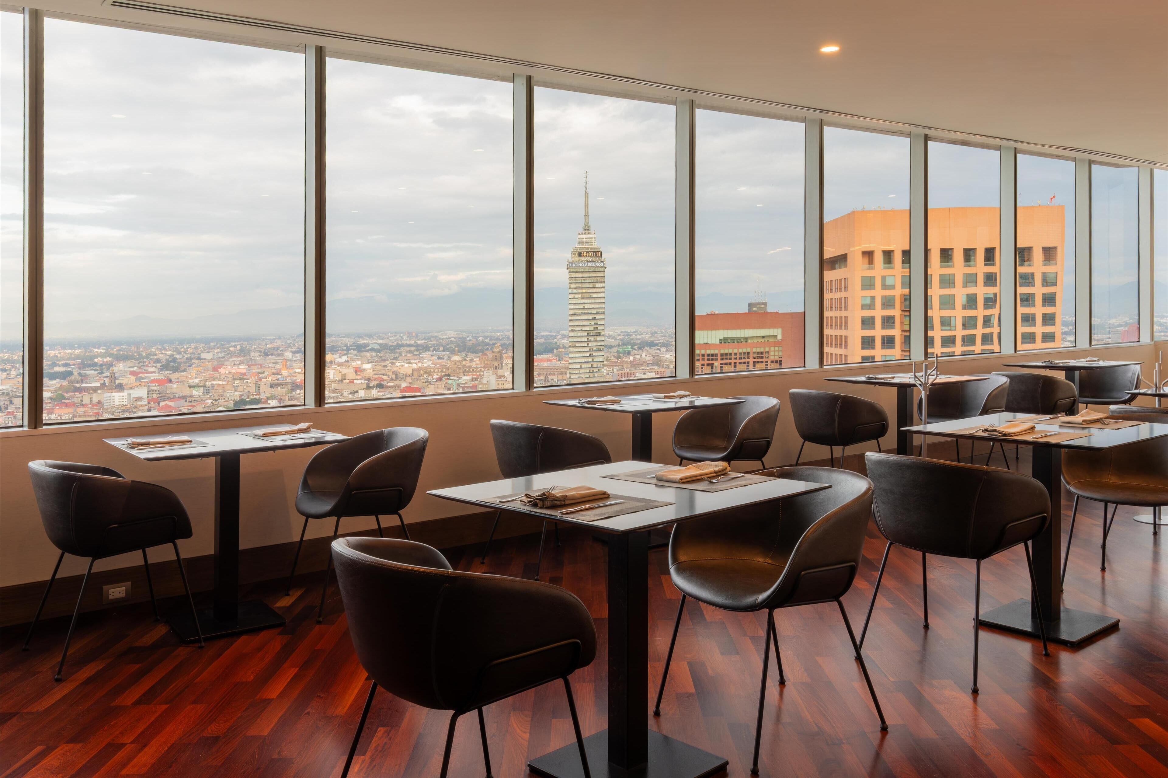 Un comedor moderno con ventanas amplias que ofrecen vista a la ciudad, incluyendo una torre destacada. Las mesas están dispuestas con sillas y el suelo es de madera.