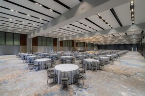 Un amplio salón de conferencias con mesas y sillas redondas, techos altos con iluminación empotrada y grandes ventanales.