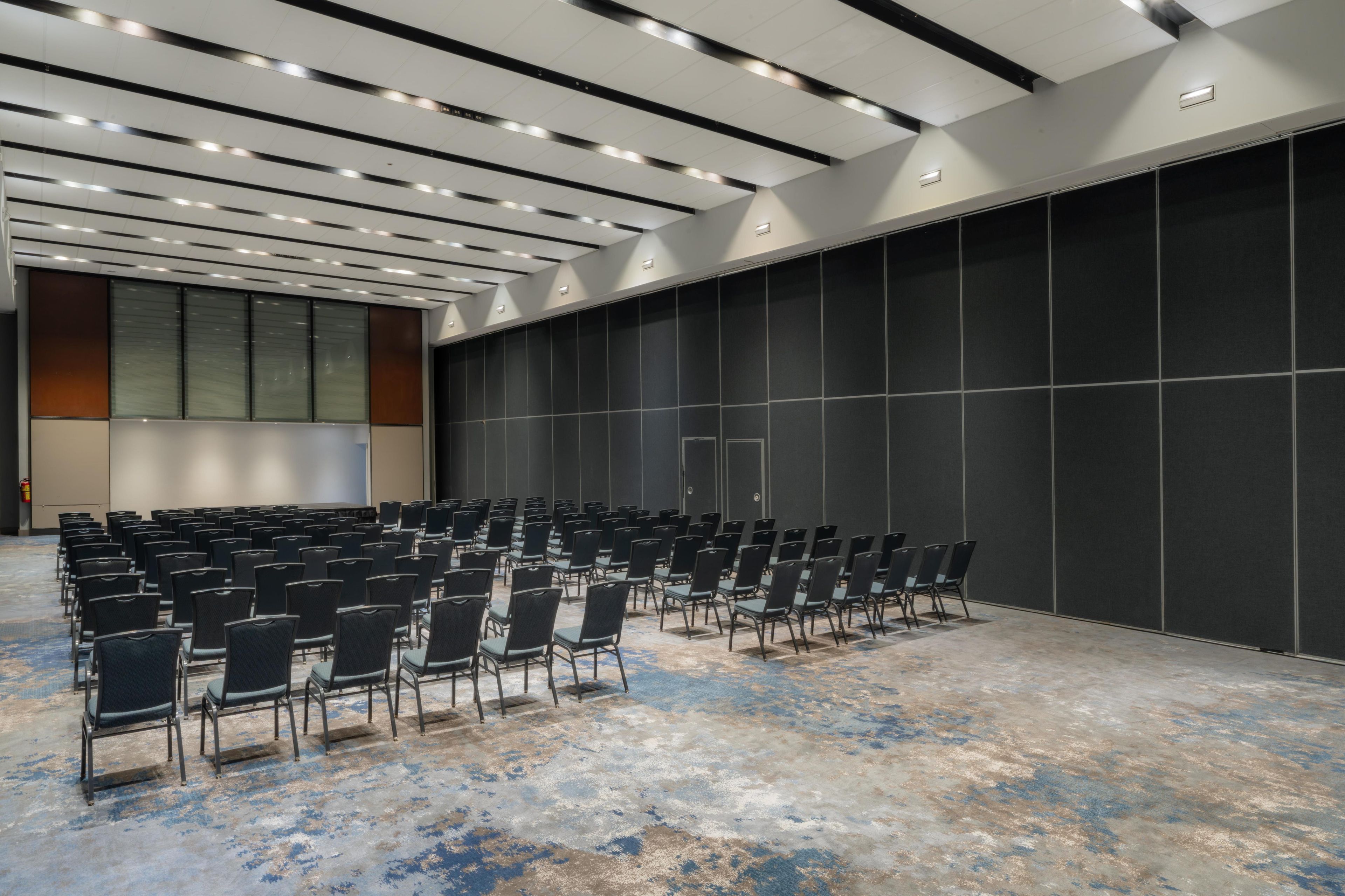 Une salle de conférence avec des rangées de chaises, un grand mur noir et un plafond doté d’un éclairage encastré.