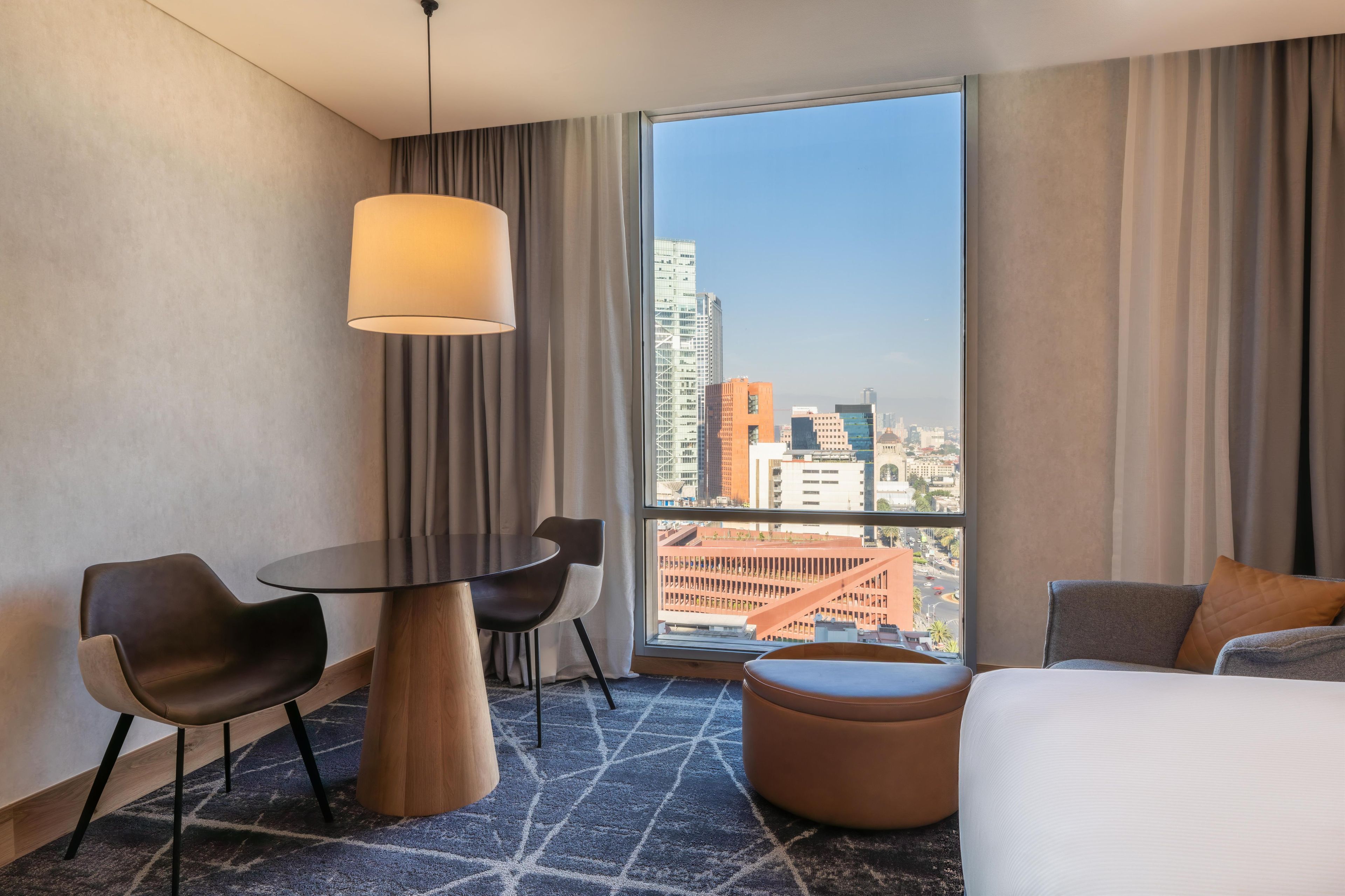 A hotel room with a large window, a round table, two chairs, a sofa, and a hanging lamp.