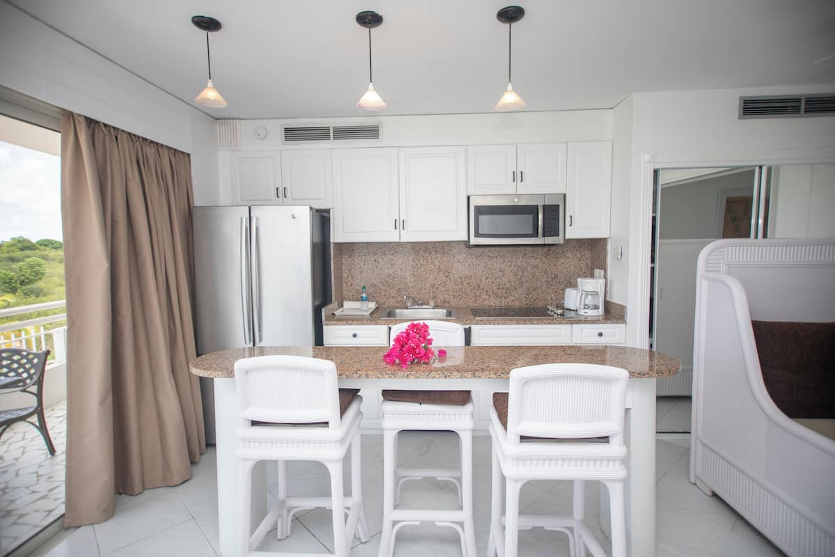 A modern kitchen with white cabinets, a granite countertop, a stainless steel refrigerator, a microwave, and a coffee maker. There are three white bar stools and a pink flower on the counter.