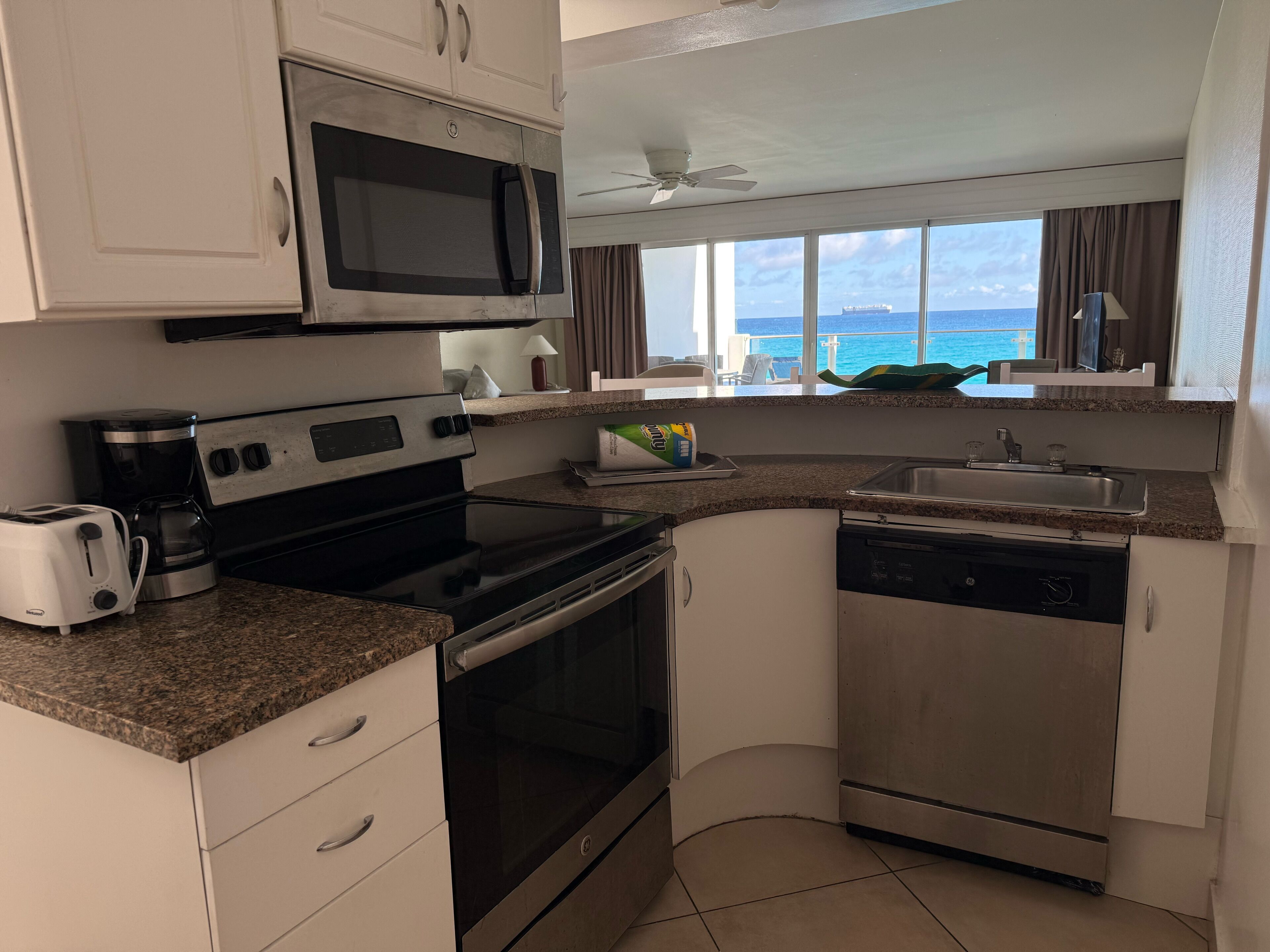 A compact kitchen with a microwave, stove, toaster, and dishwasher. It features a window with a view of the ocean and a ceiling fan.