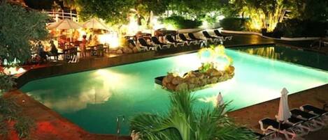 Un área de piscina nocturna con sillones reclinables y sombrillas, rodeada de árboles y un patio bien iluminado.