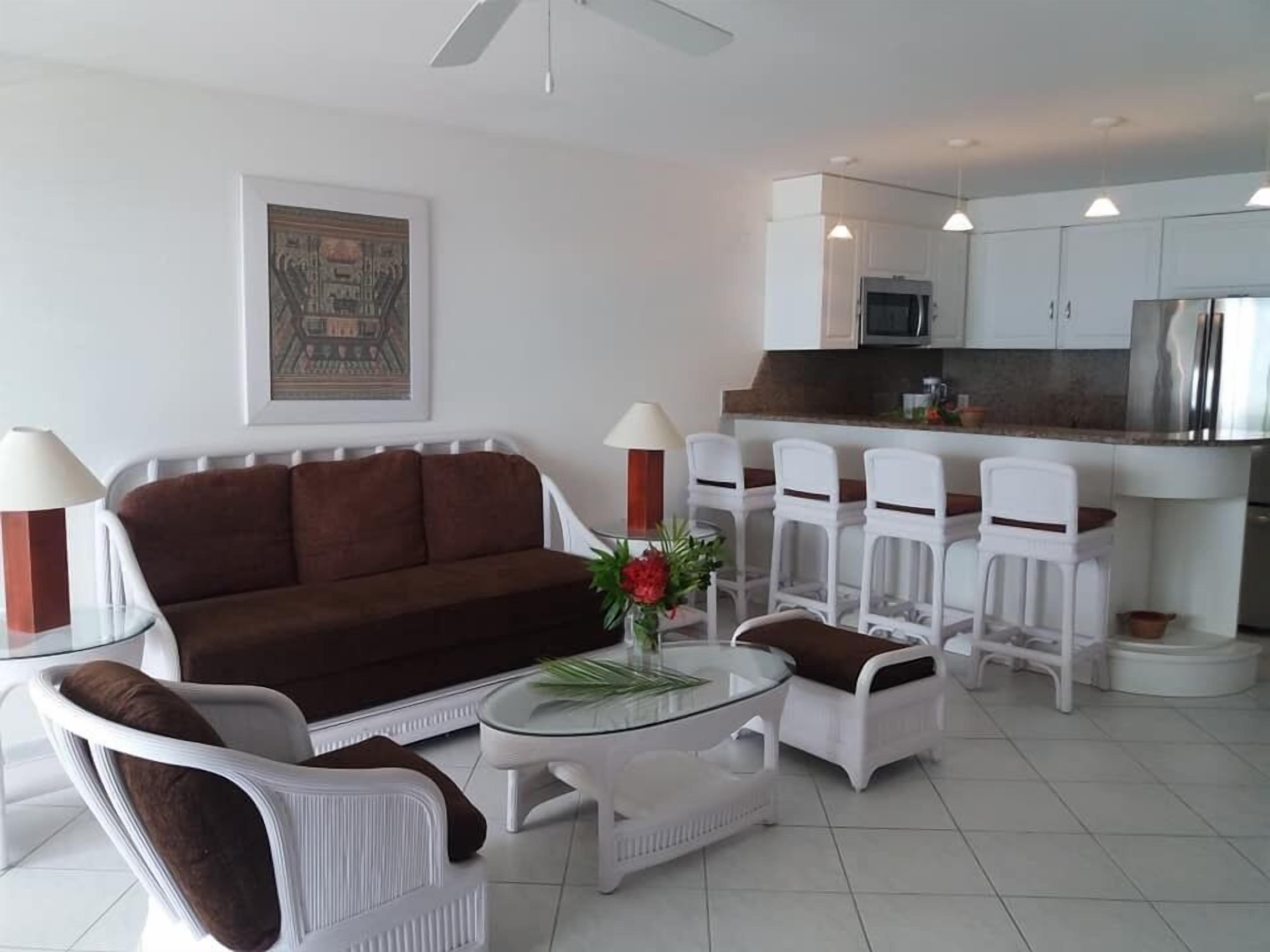 Uma sala de estar com um sofá marrom, cadeiras brancas, uma mesa de centro de vidro e uma cozinha ao fundo.