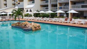 A swimming pool with lounge chairs and umbrellas, surrounded by a multi-story building with balconies.