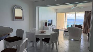 Amplia habitación con mesa de comedor, sillas blancas, ventilador de techo y vista al océano a través de amplias ventanas.