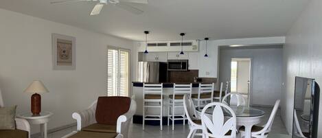 A modern living room with a dining area, white furniture, a ceiling fan, and a kitchen with a refrigerator and microwave.