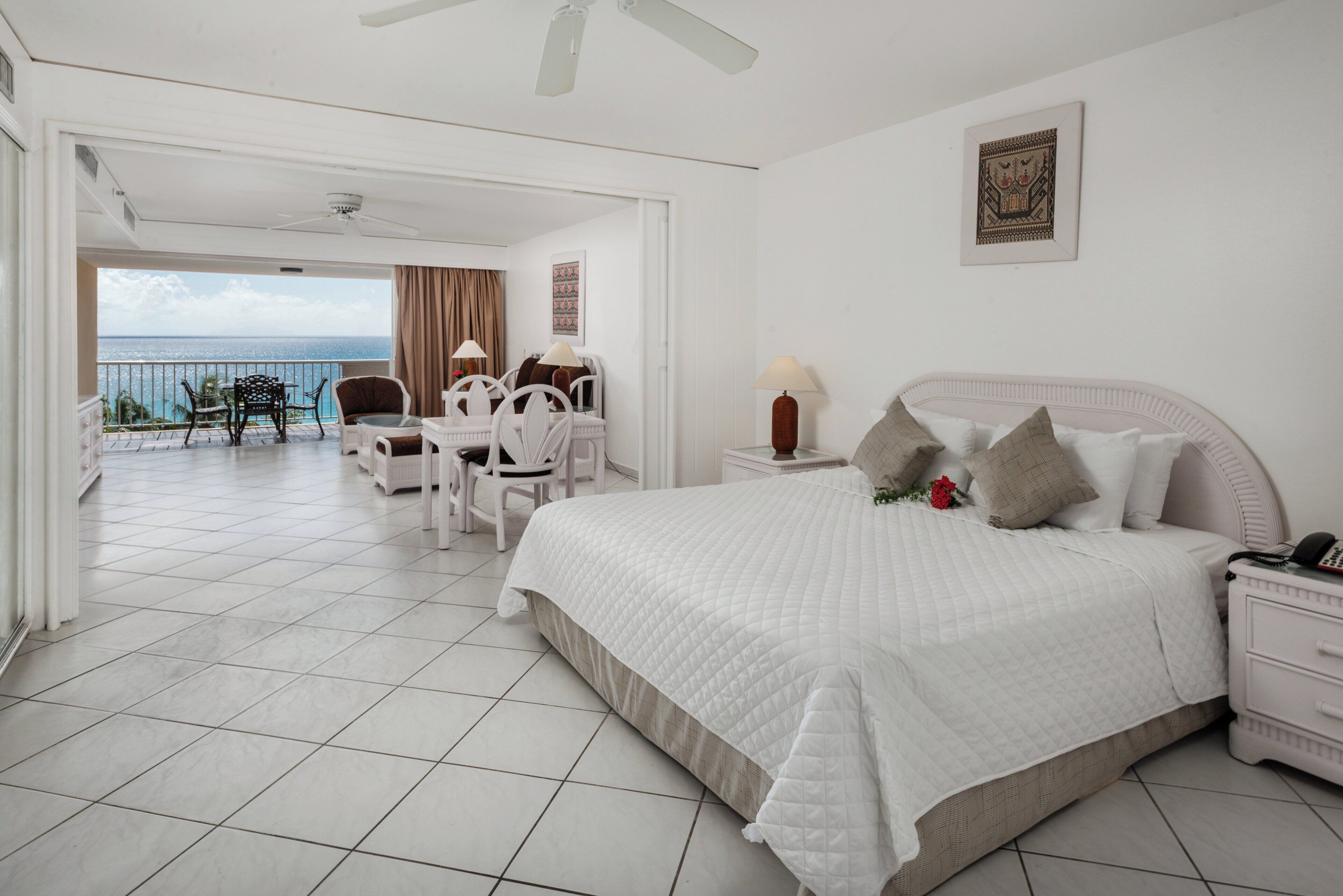 A hotel room with a large bed, a desk with a chair, a small table with a lamp, and a view of the ocean.