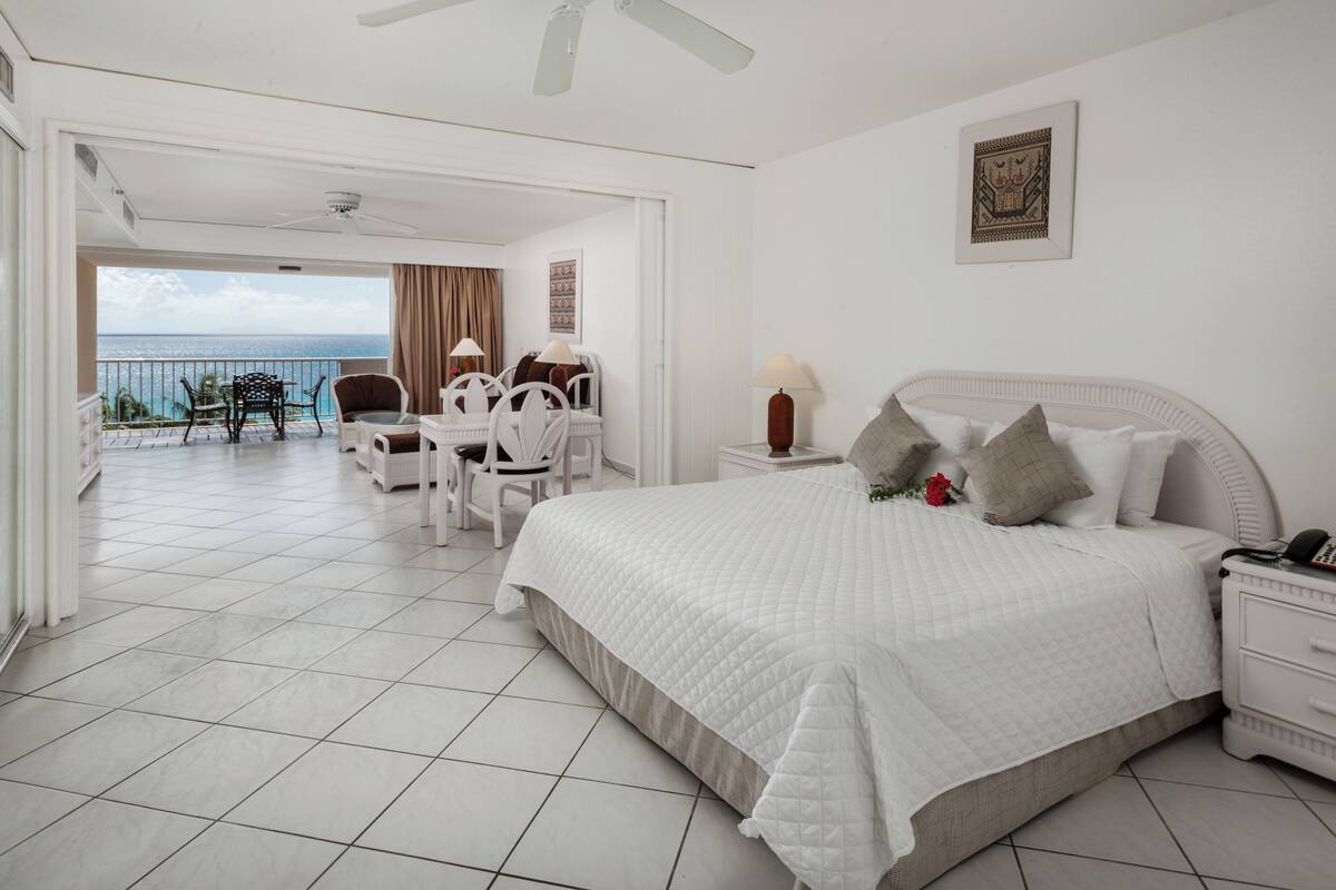 A hotel room with a large bed, a desk with a chair, a small table with a lamp, and a view of the ocean.